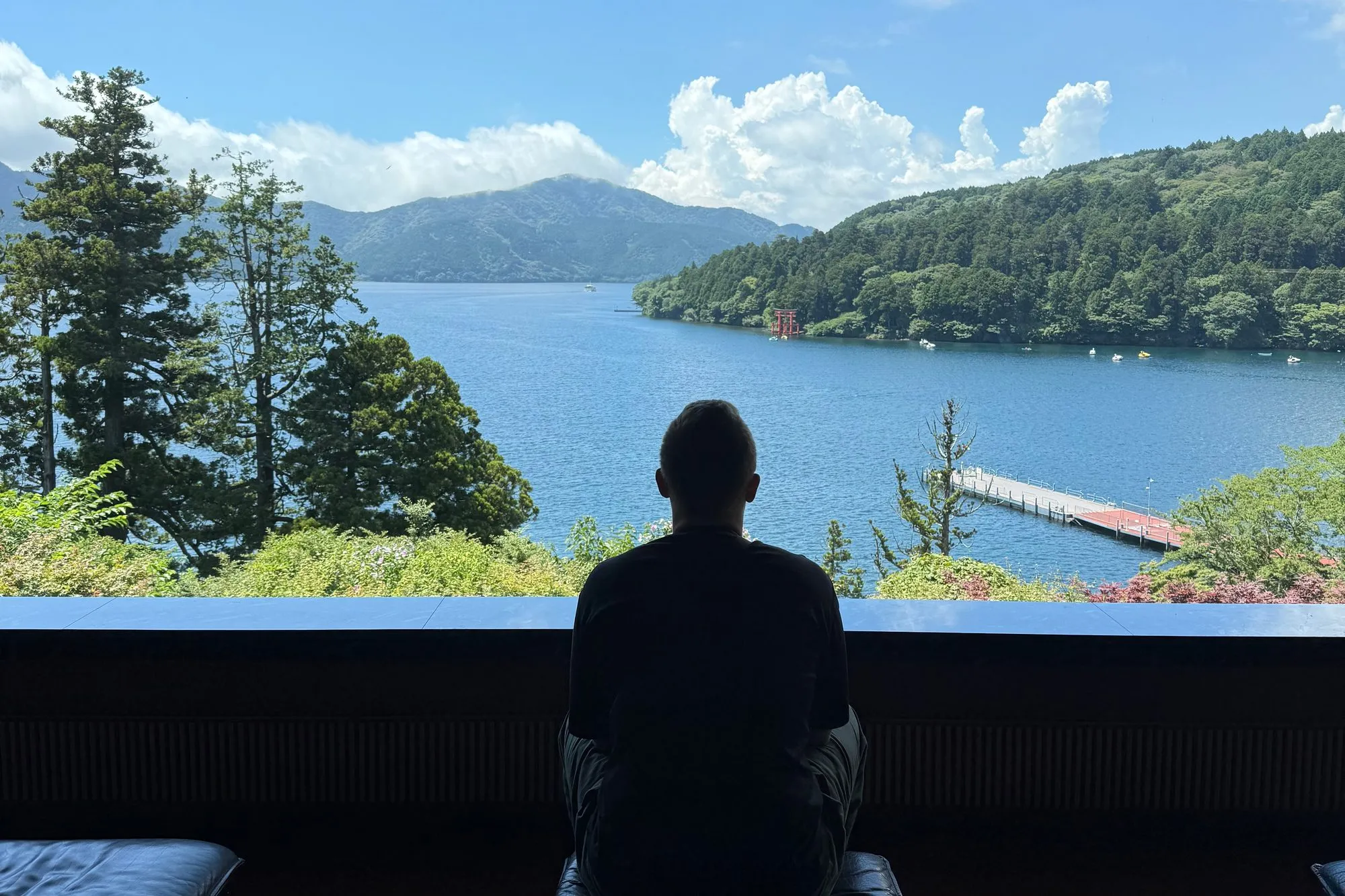 Marvin Strauch, von hinten gesehen, sitzt vor einem Panoramafenster und blickt auf einen See mit bewaldeten Bergen.